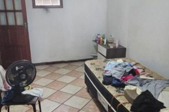 Quarto simples com roupas em cama e ventilador.