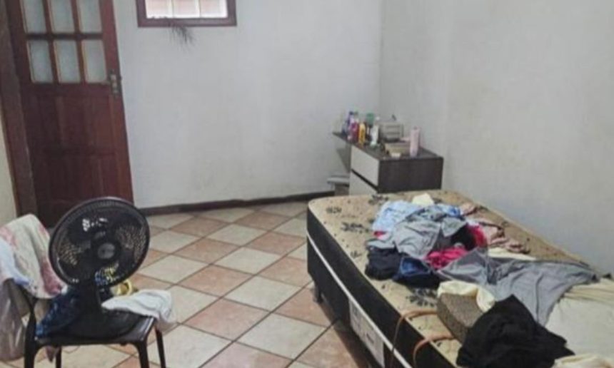 Quarto simples com roupas em cama e ventilador.