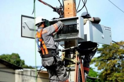 Técnicos da concessionária de energia trabalham em poste, possivelmente em manutenção ou instalação.