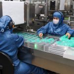 Trabalhadores em fábrica manuseando seringas em processo produtivo.