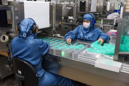 Trabalhadores em fábrica manuseando seringas em processo produtivo.