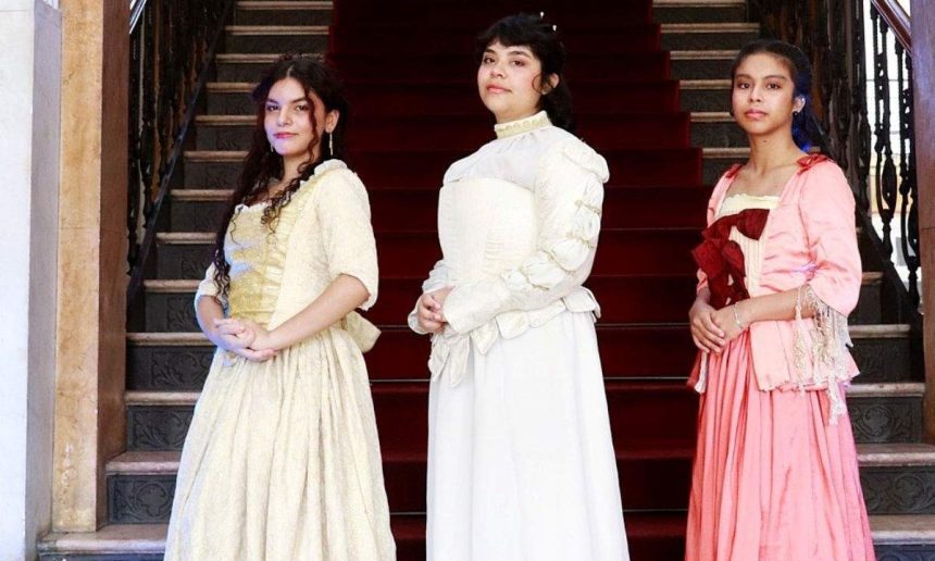 Três mulheres em trajes de época posam em frente a escadaria do Palácio da Justiça.