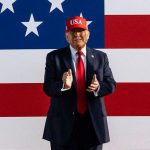Imagem colorida mostra Donald Trump com boné vermelho e bandeira dos EUA ao fundo