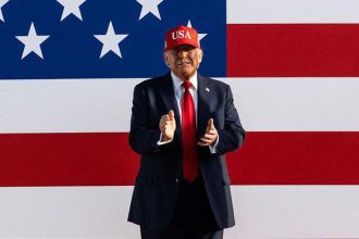 Imagem colorida mostra Donald Trump com boné vermelho e bandeira dos EUA ao fundo