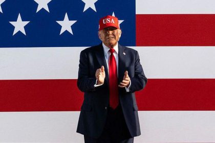 Imagem colorida mostra Donald Trump com boné vermelho e bandeira dos EUA ao fundo