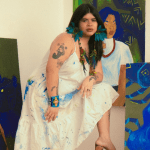 Artista Auá Mendes posando com pinturas, com vestido branco salpicado de tinta e acessórios coloridos.