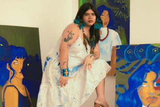 Artista Auá Mendes posando com pinturas, com vestido branco salpicado de tinta e acessórios coloridos.