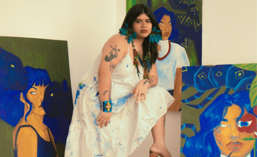 Artista Auá Mendes posando com pinturas, com vestido branco salpicado de tinta e acessórios coloridos.