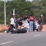 Acidente com moto em rodovia: veículo no chão, pessoas ao redor e vegetação ao fundo.
