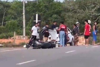 Acidente com moto em rodovia: veículo no chão, pessoas ao redor e vegetação ao fundo.
