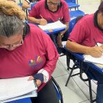 Alunos em sala de aula fazendo prova. Mulheres escrevem em cadernos e provas, usando óculos e camisetas.