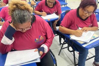 Alunos em sala de aula fazendo prova. Mulheres escrevem em cadernos e provas, usando óculos e camisetas.