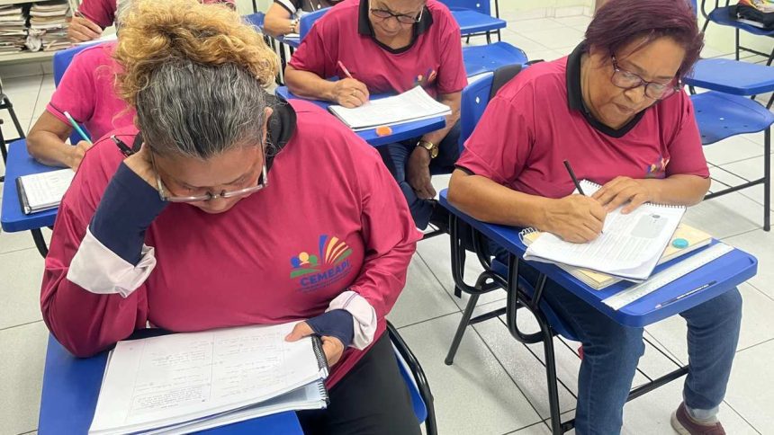 Alunos em sala de aula fazendo prova. Mulheres escrevem em cadernos e provas, usando óculos e camisetas.