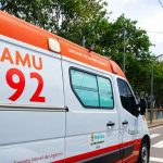 Ambulância do SAMU 192 em via pública, com sinalização de trânsito e vegetação ao fundo.