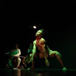 Apresentação de dança contemporânea no palco, com iluminação verde e foco no dançarino principal.