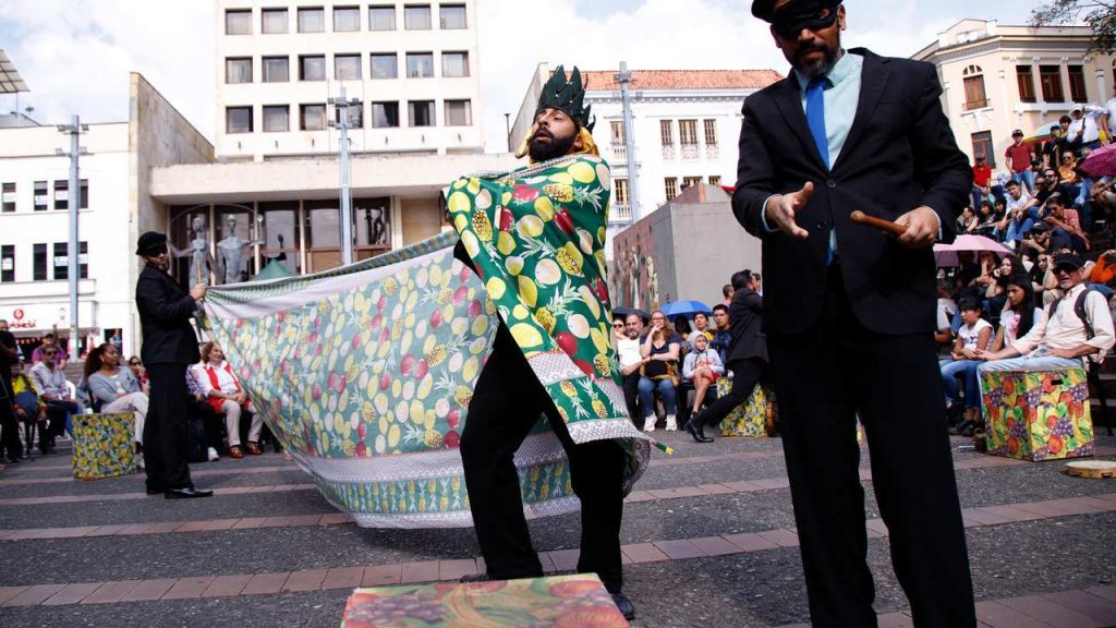 Artistas performam em evento cultural ao ar livre, com roupas coloridas e máscaras.