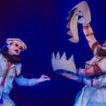 Atores em figurinos elaborados durante uma apresentação teatral com adereços de papelão.