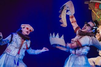 Atores em figurinos elaborados durante uma apresentação teatral com adereços de papelão.