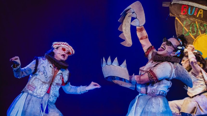 Atores em figurinos elaborados durante uma apresentação teatral com adereços de papelão.