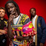Banda musical Guiss Guiss Bou Bess com três membros em estúdio, fundo vermelho. Destaque para roupas coloridas e estilosas.