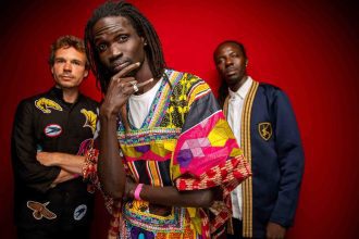 Banda musical Guiss Guiss Bou Bess com três membros em estúdio, fundo vermelho. Destaque para roupas coloridas e estilosas.