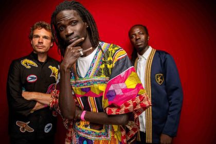 Banda musical Guiss Guiss Bou Bess com três membros em estúdio, fundo vermelho. Destaque para roupas coloridas e estilosas.