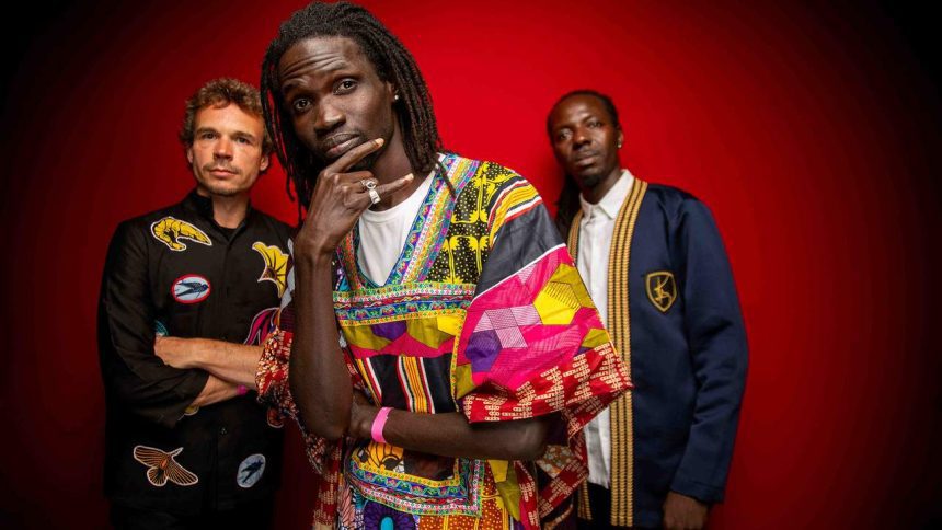 Banda musical Guiss Guiss Bou Bess com três membros em estúdio, fundo vermelho. Destaque para roupas coloridas e estilosas.