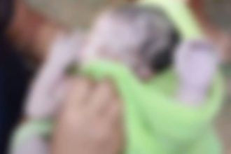 Bebê recém-nascido envolto em cobertor verde, sendo segurado por alguém.