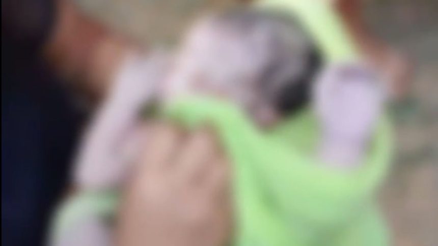 Bebê recém-nascido envolto em cobertor verde, sendo segurado por alguém.