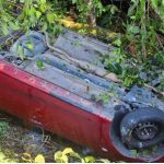Carro vermelho capotado em meio à vegetação após acidente.