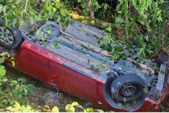 Carro vermelho capotado em meio à vegetação após acidente.