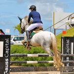 Cavaleira montando a cavalo branco saltando um obstáculo em competição de hipismo.