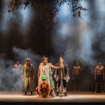 Cena teatral com personagens em figurinos coloridos, representando a mitologia Tukano.