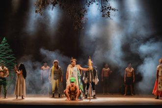 Cena teatral com personagens em figurinos coloridos, representando a mitologia Tukano.