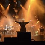 Concerto de orquestra e banda de New Metal no palco, com maestro e músicos.
