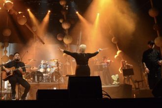 Concerto de orquestra e banda de New Metal no palco, com maestro e músicos.