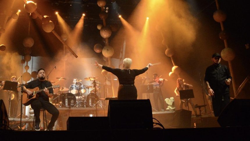 Concerto de orquestra e banda de New Metal no palco, com maestro e músicos.
