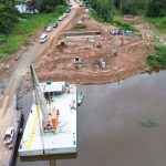 Construção de ponte sobre rio na BR-319, com balsas, guindaste e veículos na área.