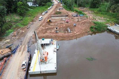 Construção de ponte sobre rio na BR-319, com balsas, guindaste e veículos na área.