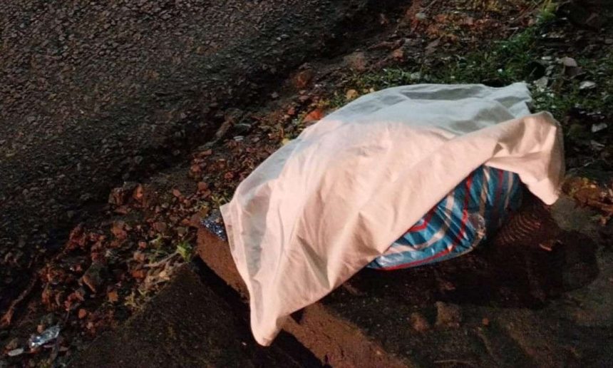 Corpo de bebê coberto por tecido branco na rua, próximo a vegetação e asfalto.