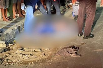 Corpo de homem coberto na rua, cena de crime com pessoas ao redor.