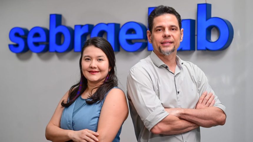 Dois profissionais sorrindo em frente ao logo da Sebrae, com inscrições para capacitação de artistas visuais amazônicos.