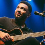 Eli Soares tocando violão em show, com microfone e fundo azul