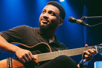 Eli Soares tocando violão em show, com microfone e fundo azul