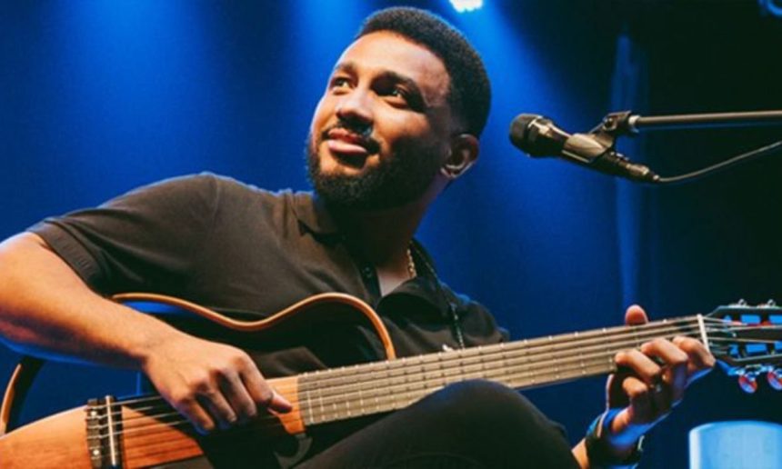 Eli Soares tocando violão em show, com microfone e fundo azul