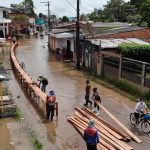 Enchente em área urbana, com pessoas construindo passarela de madeira para travessia.