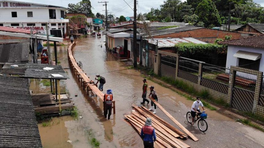Enchente em área urbana, com pessoas construindo passarela de madeira para travessia.