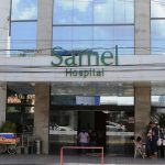 Fachada do Hospital Samel com entrada e letreiro verde. Pessoas em frente.