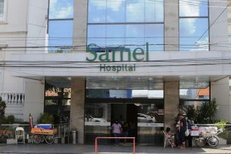 Fachada do Hospital Samel com entrada e letreiro verde. Pessoas em frente.