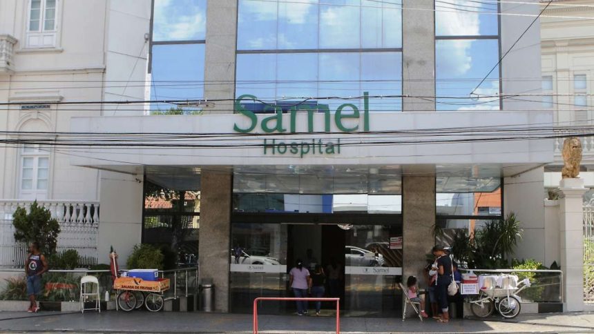 Fachada do Hospital Samel com entrada e letreiro verde. Pessoas em frente.